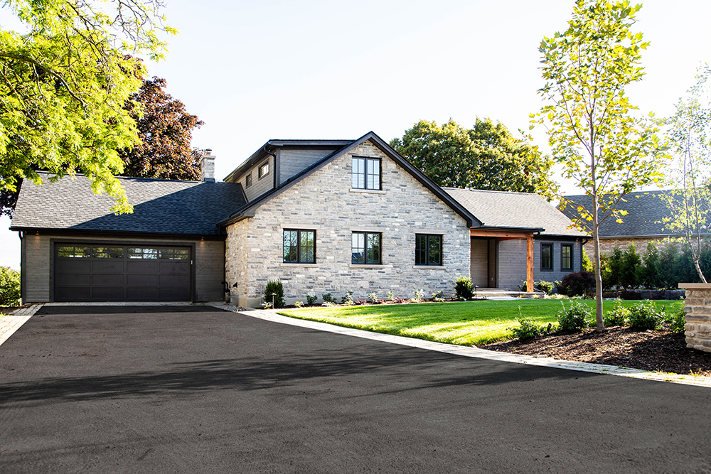 A modern single-story house with a stone facade, black roof, and an attached two-car garage, surrounded by a well-manicured lawn and young trees.