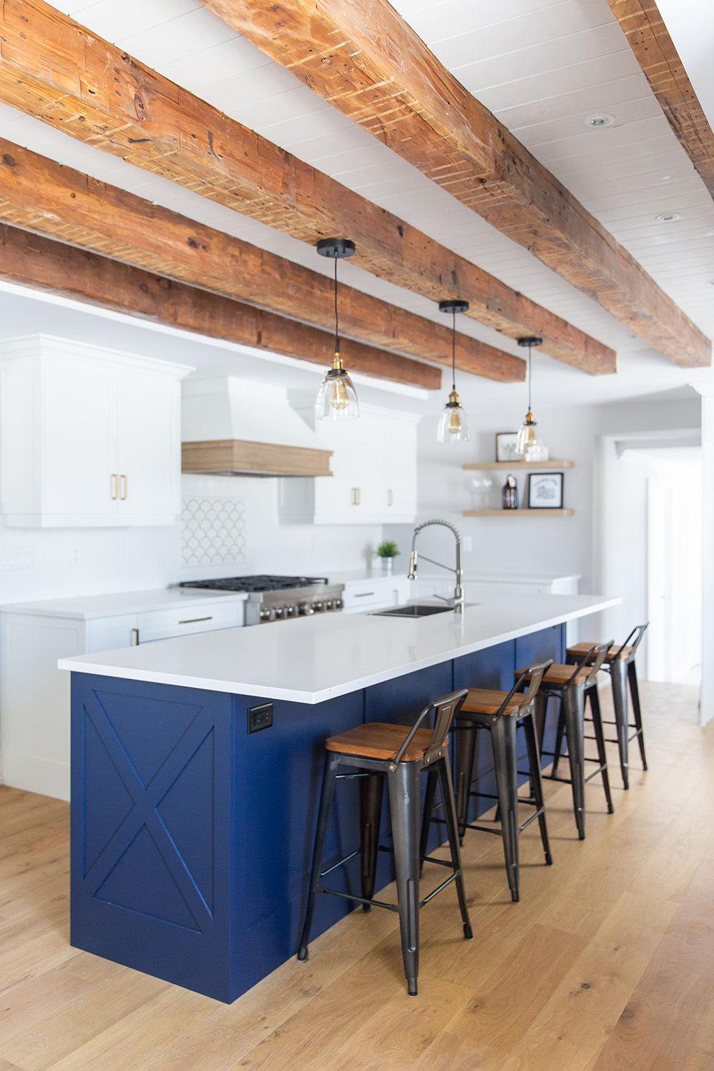 A modern kitchen with white cabinets, a blue island, wooden floors, and exposed ceiling beams, with pendant lights and bar stools.