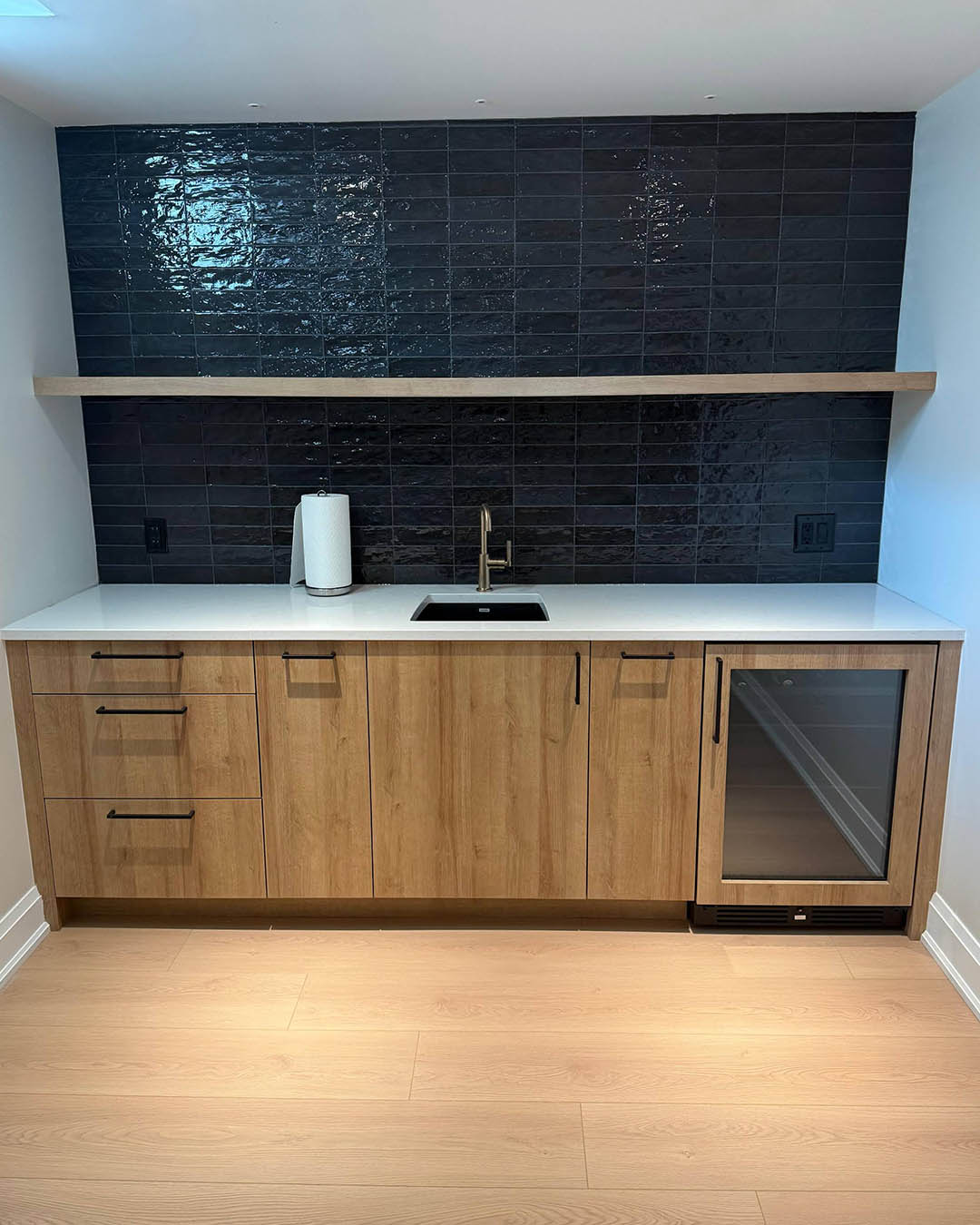 Modern kitchen cabinetry with wood finish, white countertop, black backsplash tiles, sink, and integrated wine cooler, showcasing Heartwood Renovations' design expertise in home renovations.