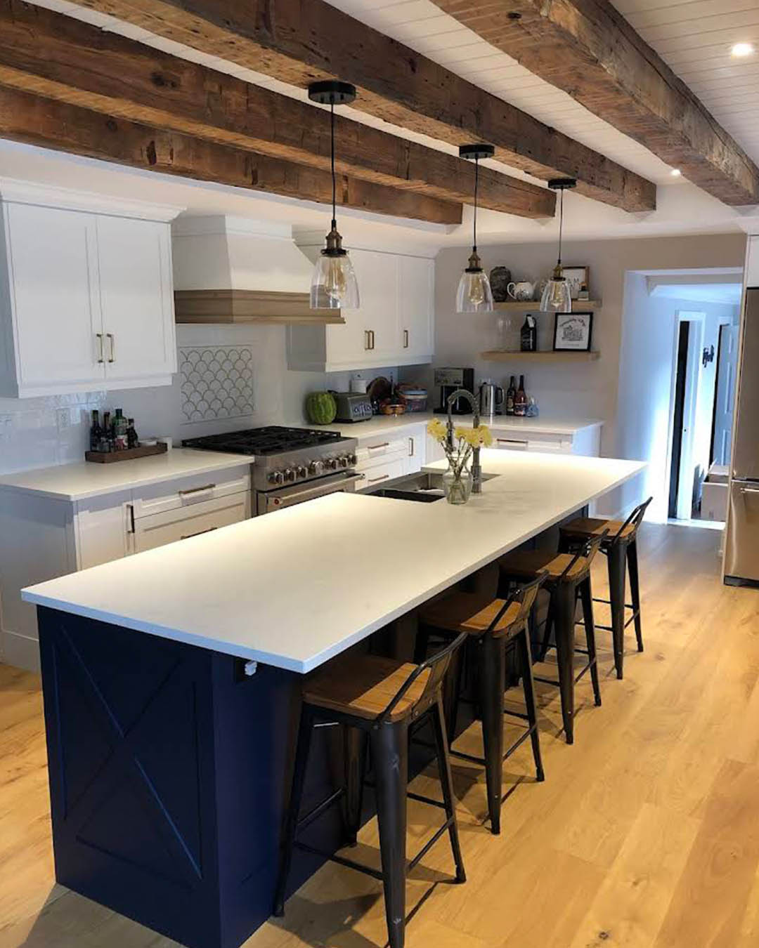 Modern kitchen featuring a large white island with dark blue cabinetry, wooden bar stools, exposed wooden beams, and stainless steel appliances, showcasing Heartwood Renovations' craftsmanship and design expertise.