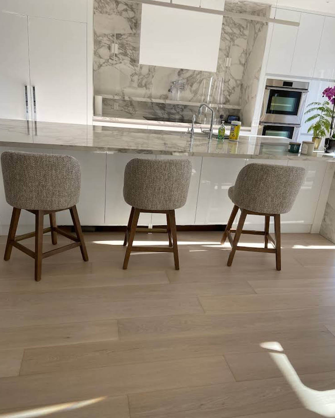 Modern kitchen with marble countertops, three upholstered bar stools, and sleek cabinetry, showcasing a sophisticated renovation by Heartwood Renovations.